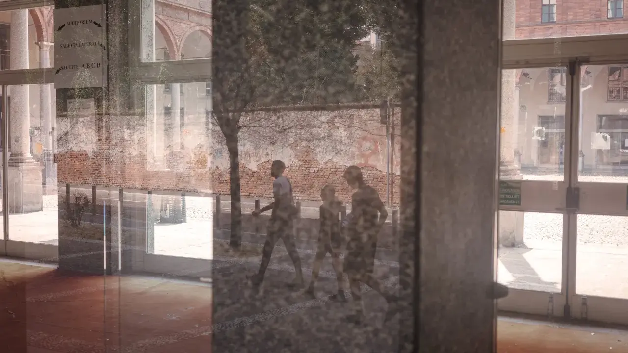 Un riflesso su vetro mostra tre figure che camminano, con un muro di mattoni e un edificio sullo sfondo.