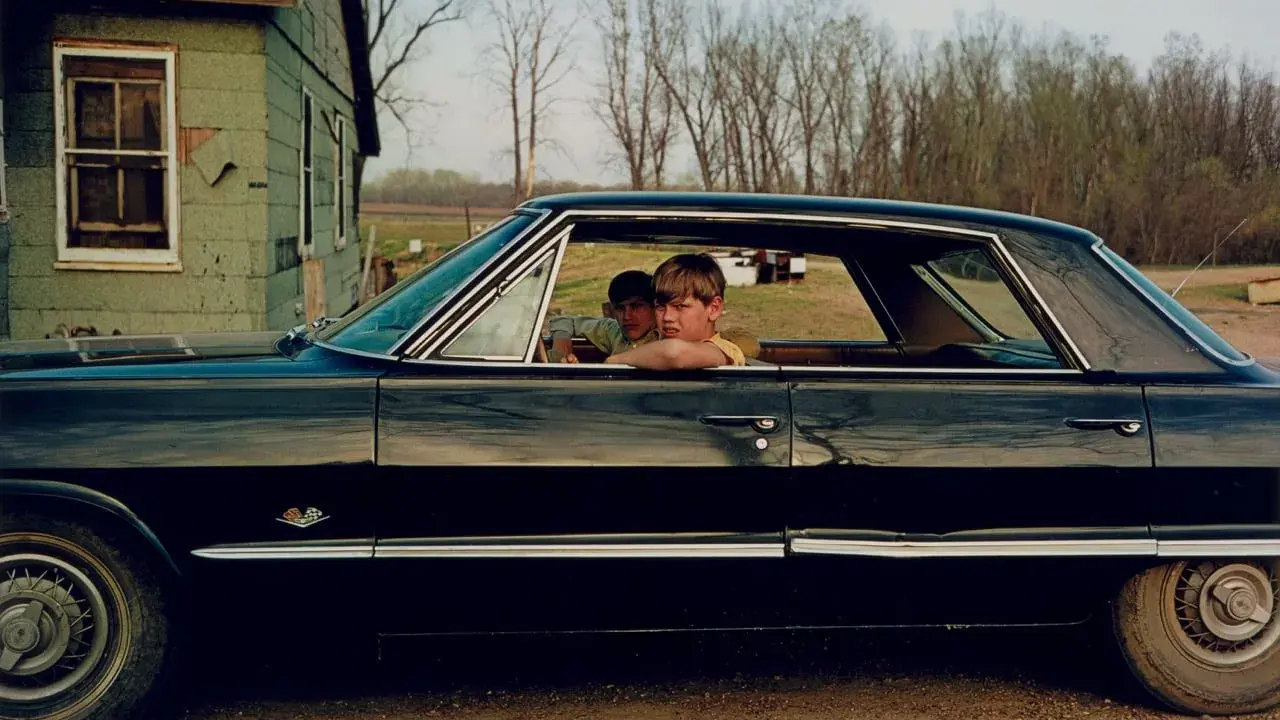 William Eggleston foto: due ragazzi guardano fuori da un'auto nera lucida, con una casa verde sullo sfondo.