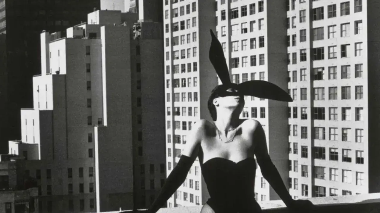 Iconica immagine di Helmut Newton: una donna con orecchie da coniglio e un corsetto nero posa su un tetto, con grattacieli sullo sfondo.
