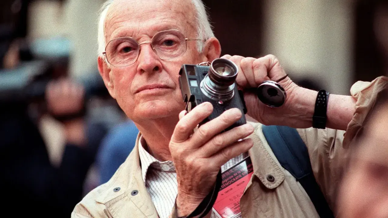 Un anziano fotografo francese famoso con occhiali tiene una vecchia macchina fotografica, pronto a catturare un momento.