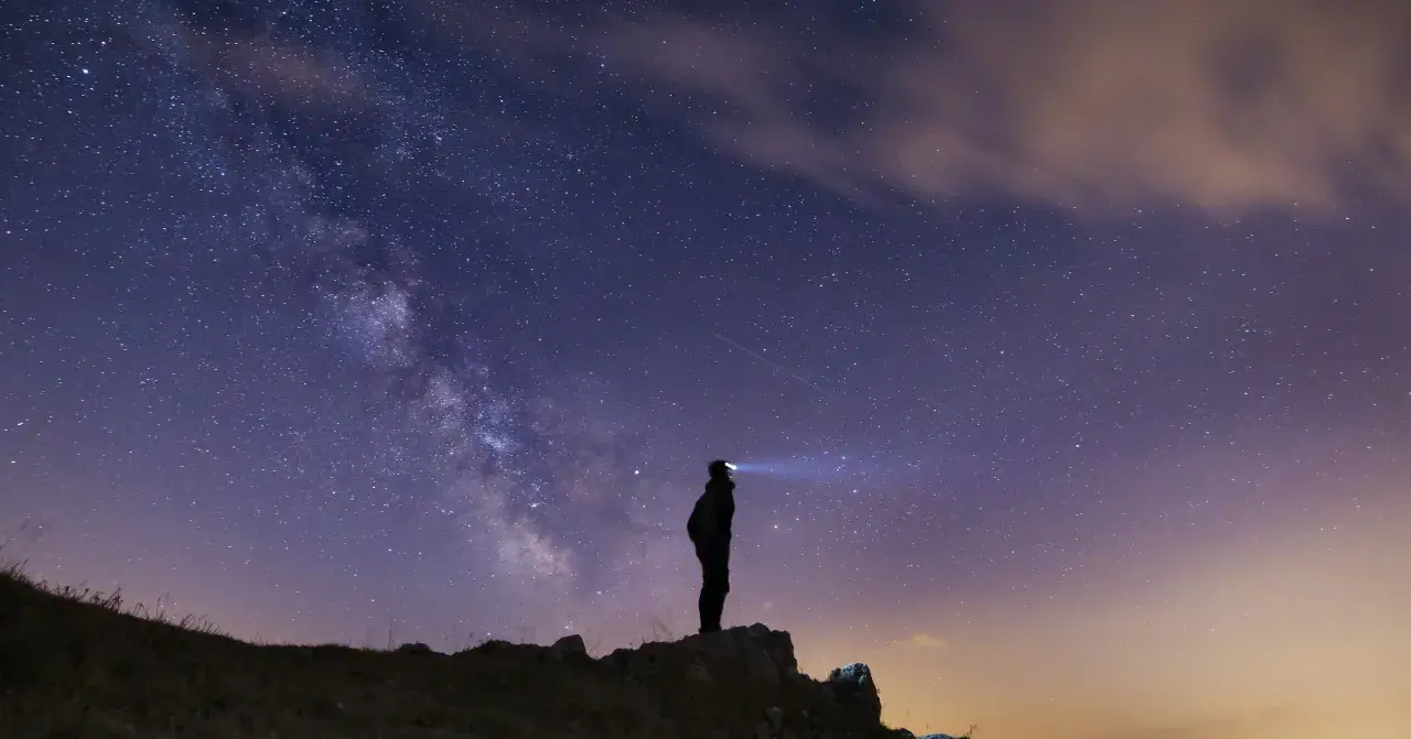 Silhouette di una persona su una collina, che guarda la Via Lattea. Un faro illumina il cielo stellato, un invito a scoprire la Via Lattea.