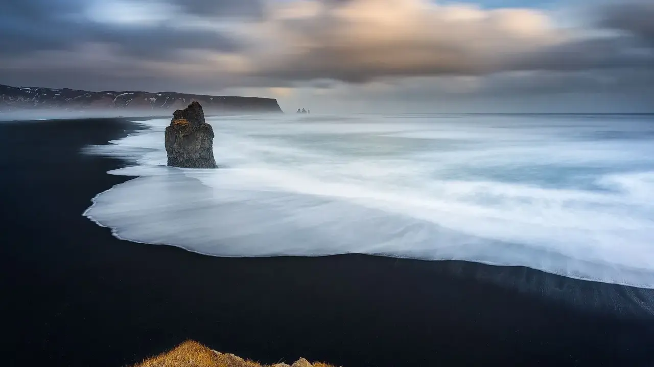 Scogliera solitaria su spiaggia nera, onde bianche in movimento con messa a fuoco infinito. Cielo drammatico.