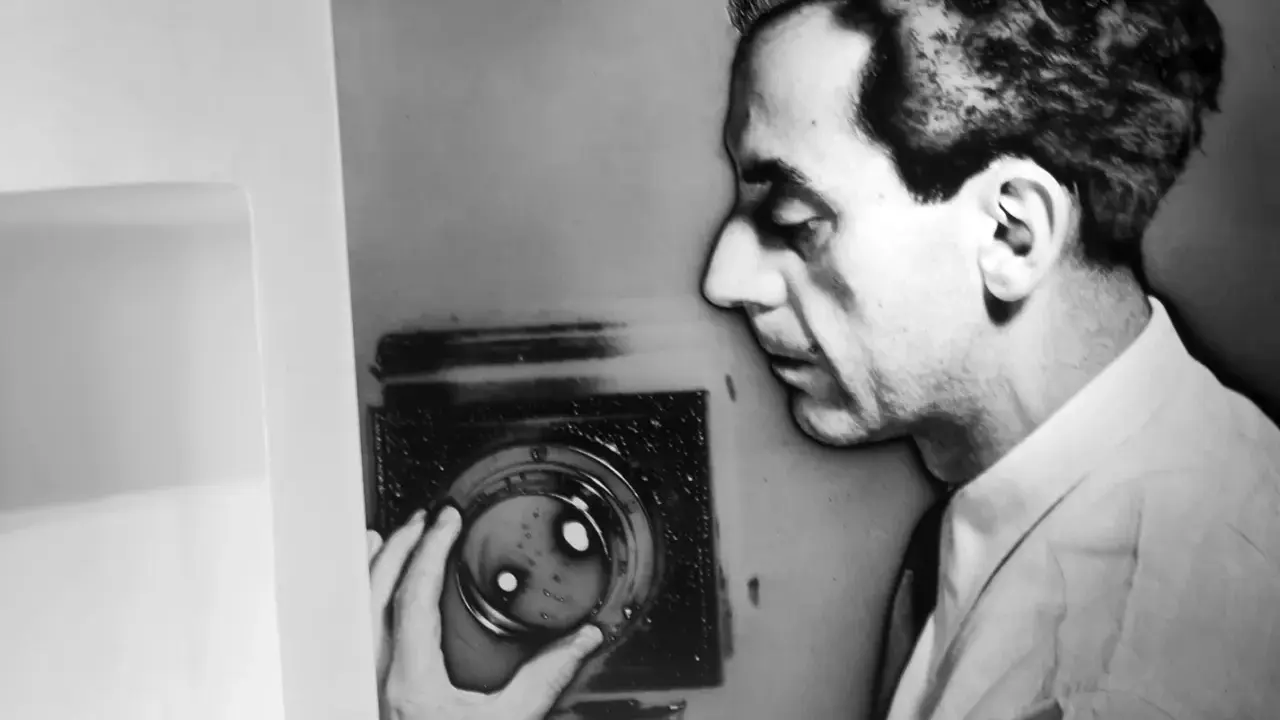 Man Ray, con la sua mano, manipola un oggetto che ricorda i suoi celebri rayogrammi, creando un'ombra enigmatica.
