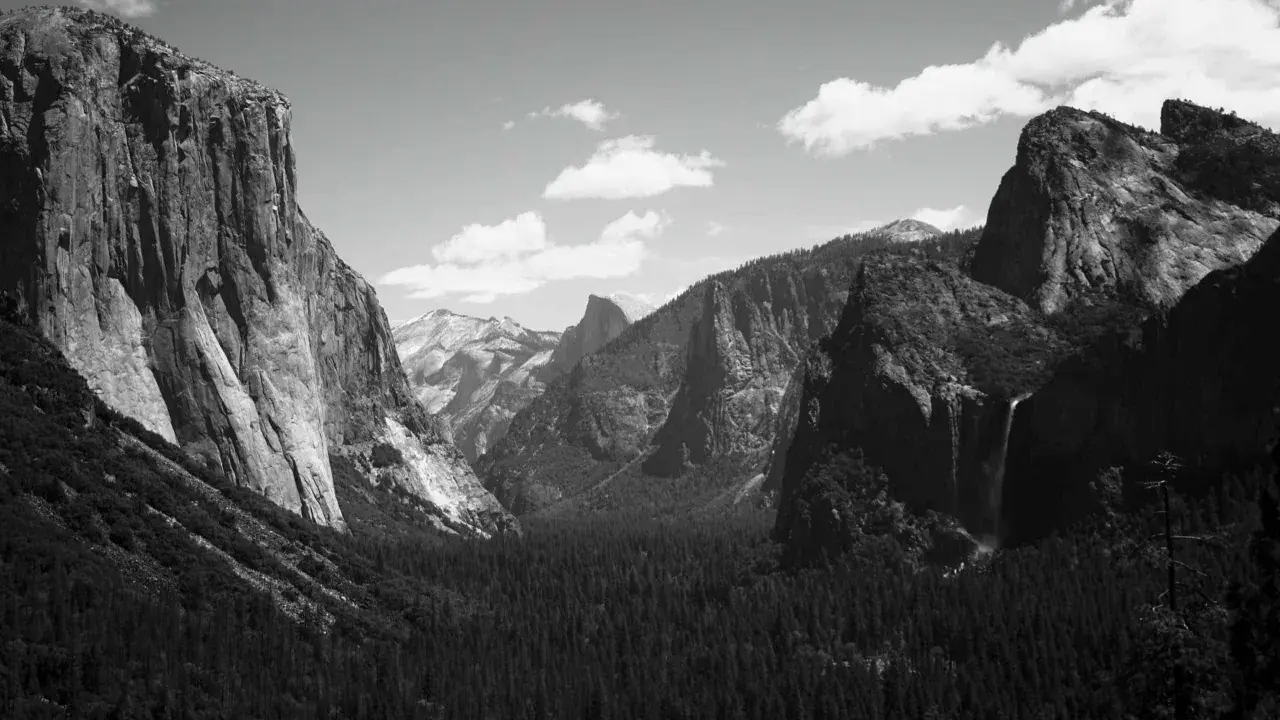 Iconici paesaggi di Yosemite, immortalati da fotografi famosi. Maestose pareti rocciose, una cascata e una fitta foresta sotto un cielo nuvoloso.