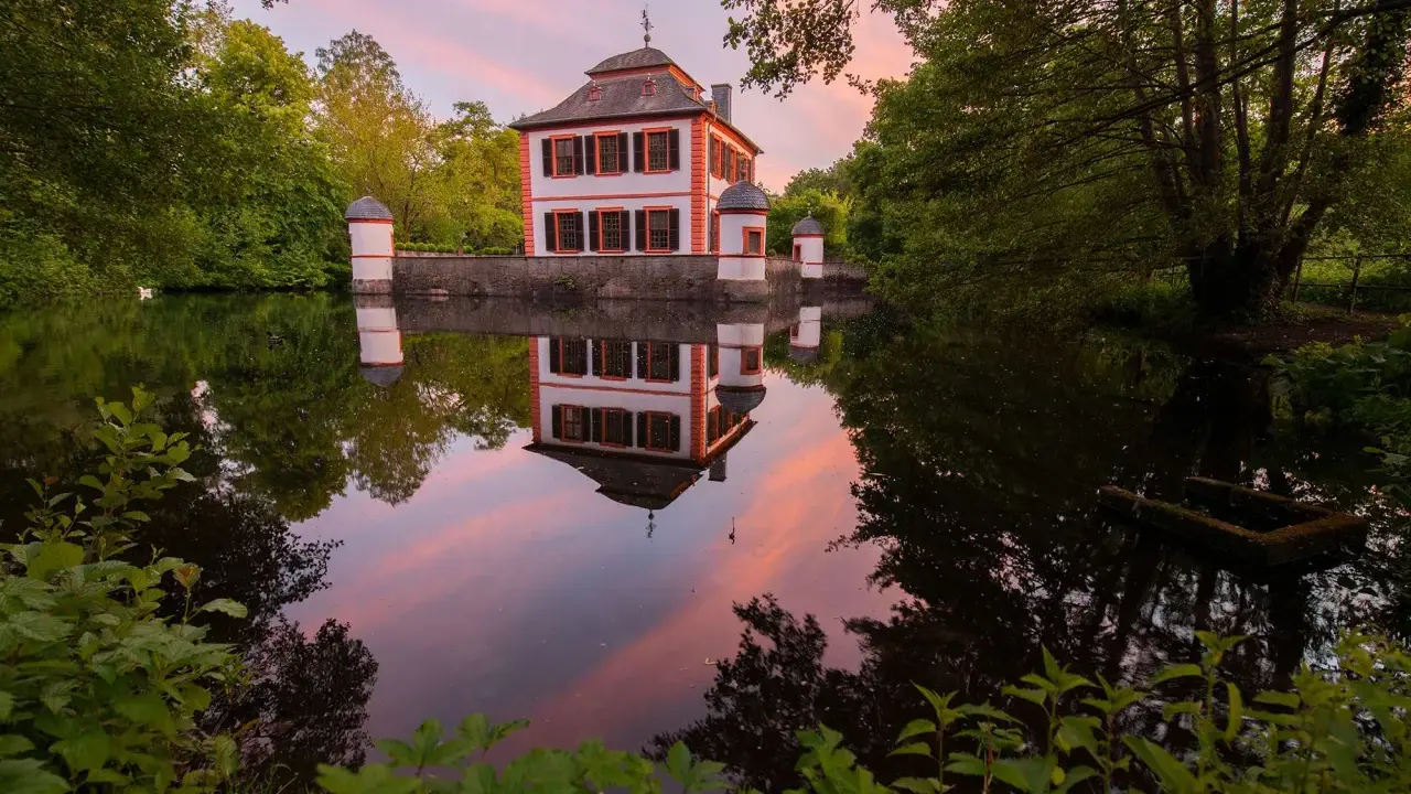 Castello bianco e arancione con torrette, riflessi nell'acqua calma al tramonto, circondato da alberi verdi.