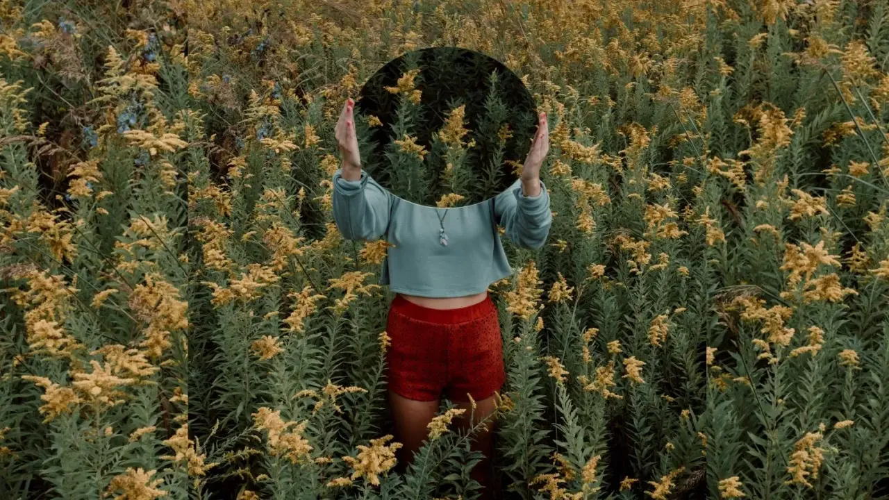 Una persona in un campo di fiori gialli tiene uno specchio rotondo davanti al viso, creando una **fotografia surrealista** dove il riflesso mostra solo la vegetazione circostante.