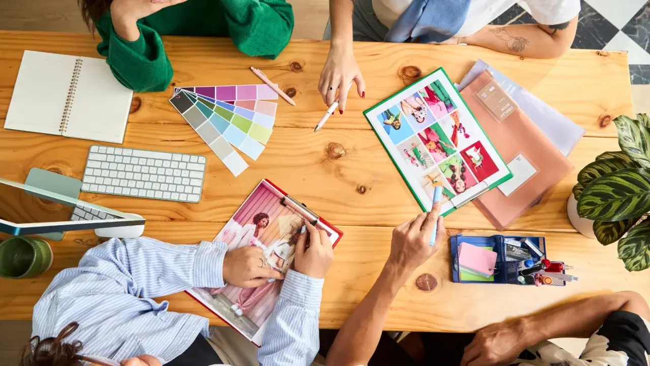 Un team creativo discute idee su un tavolo di legno, con campioni di colore, tastiera, quaderno e foto. Questo è un esempio di come una foto formato JPG può catturare un momento di collaborazione.