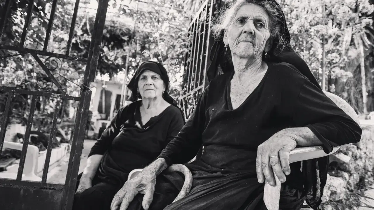 Due anziane donne, ritratte con la sensibilità di Paul Strand, siedono all'aperto, con sguardi intensi e mani segnate dal tempo.