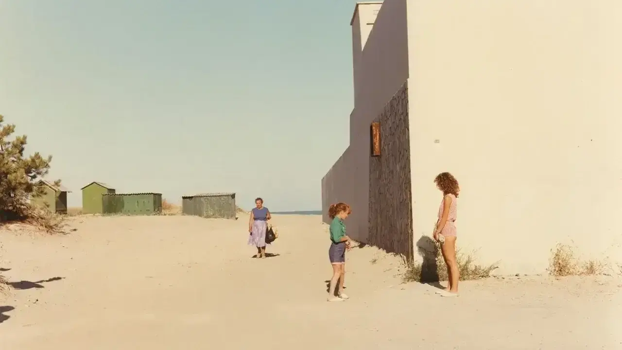 Ghirri fotografo cattura un momento estivo: due bambine e una donna sulla spiaggia, con capanne verdi sullo sfondo e un muro bianco imponente.