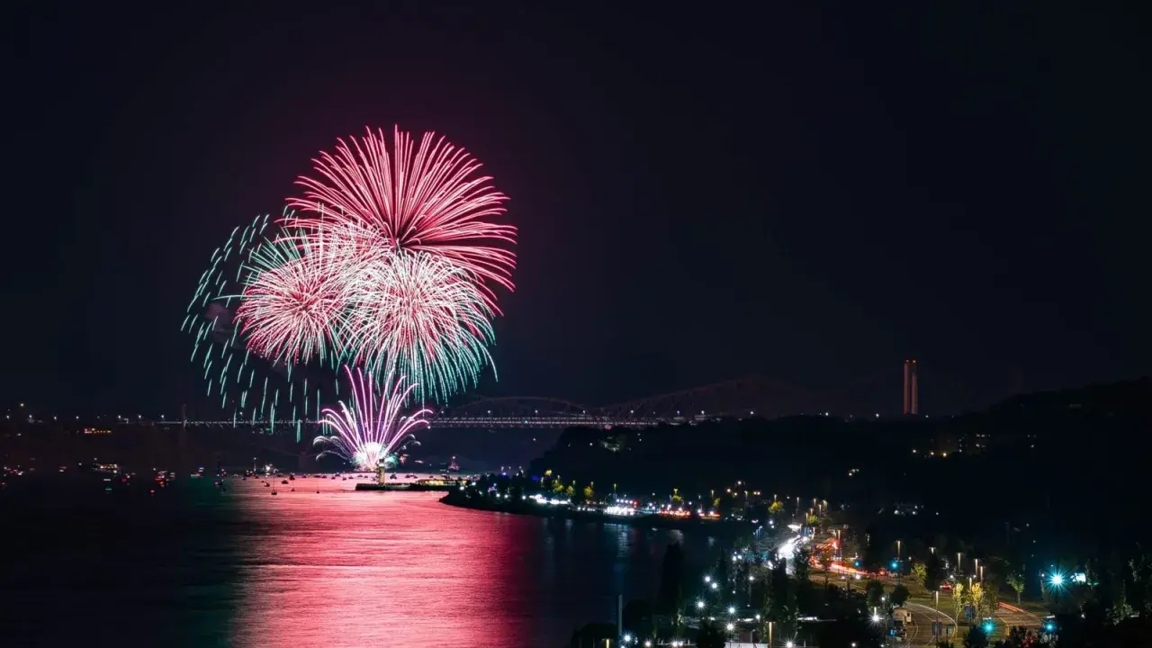 Spettacolare foto fuochi d'artificio rossi e verdi illuminano il cielo notturno sopra un fiume e una città con ponti illuminati.