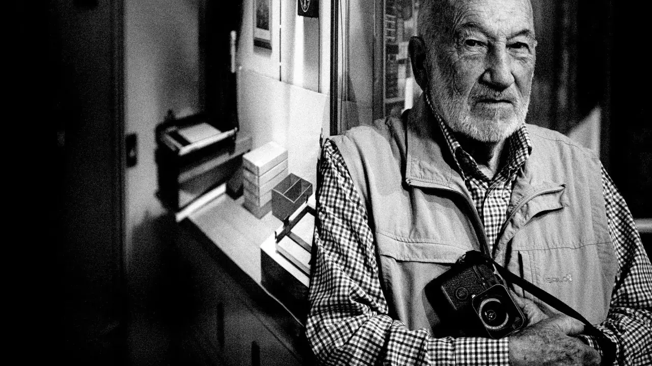 Ritratto di Gianni Berengo Gardin, con la sua inseparabile macchina fotografica al collo, sguardo intenso.