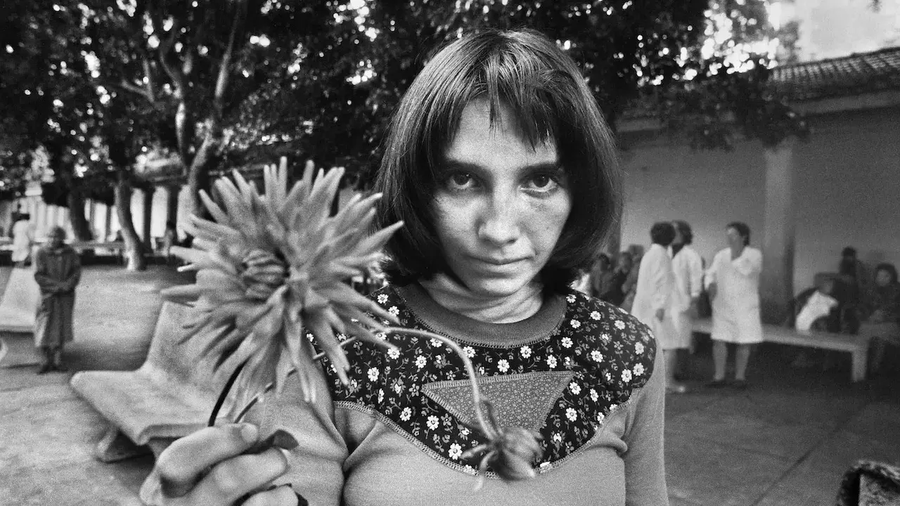 Letizia Battaglia giovane, con uno sguardo intenso, tiene un fiore. Sullo sfondo, figure sfocate in un cortile.