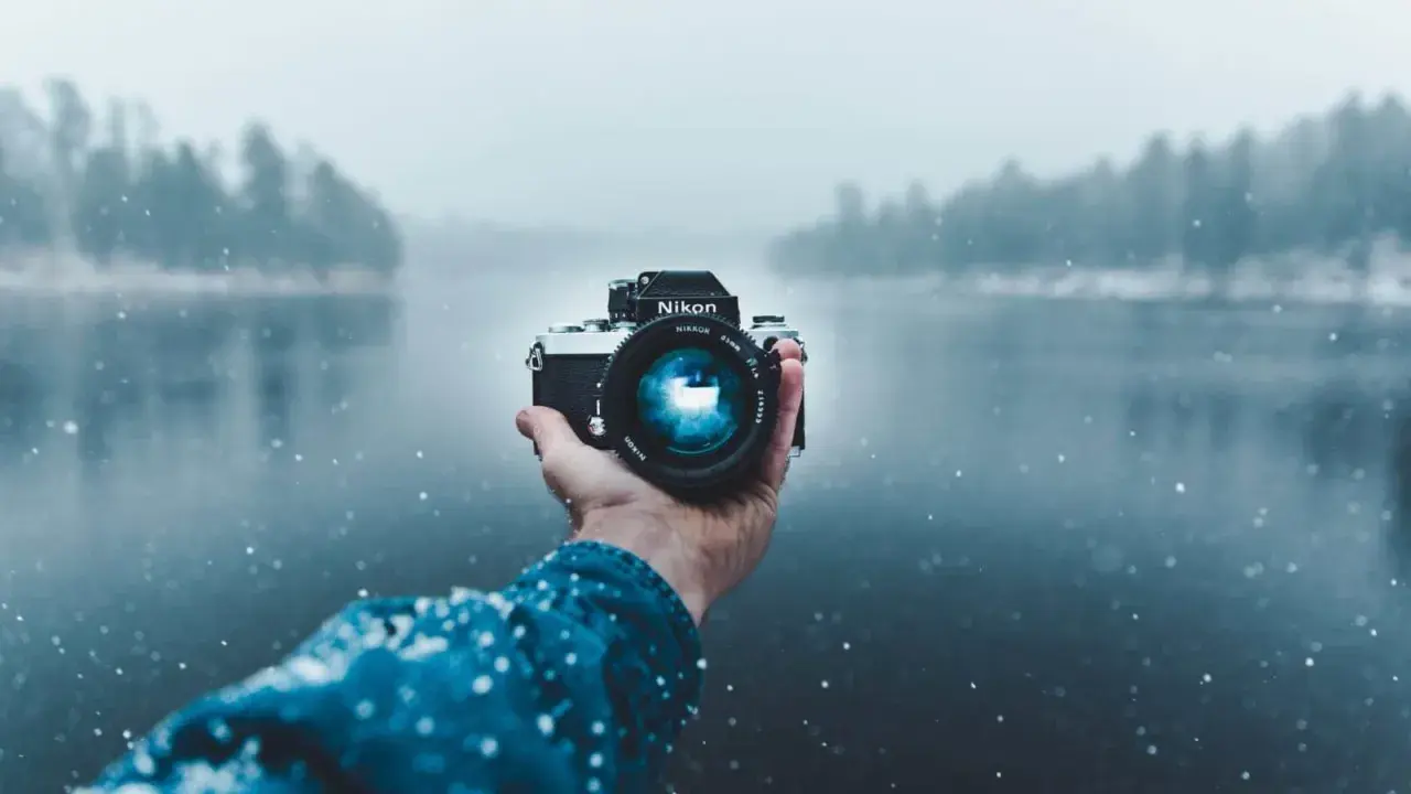 Mano che tiene una fotocamera Nikon in una giornata nevosa, con un lago e alberi sfocati sullo sfondo. Una foto raw per catturare l'essenza dell'inverno.