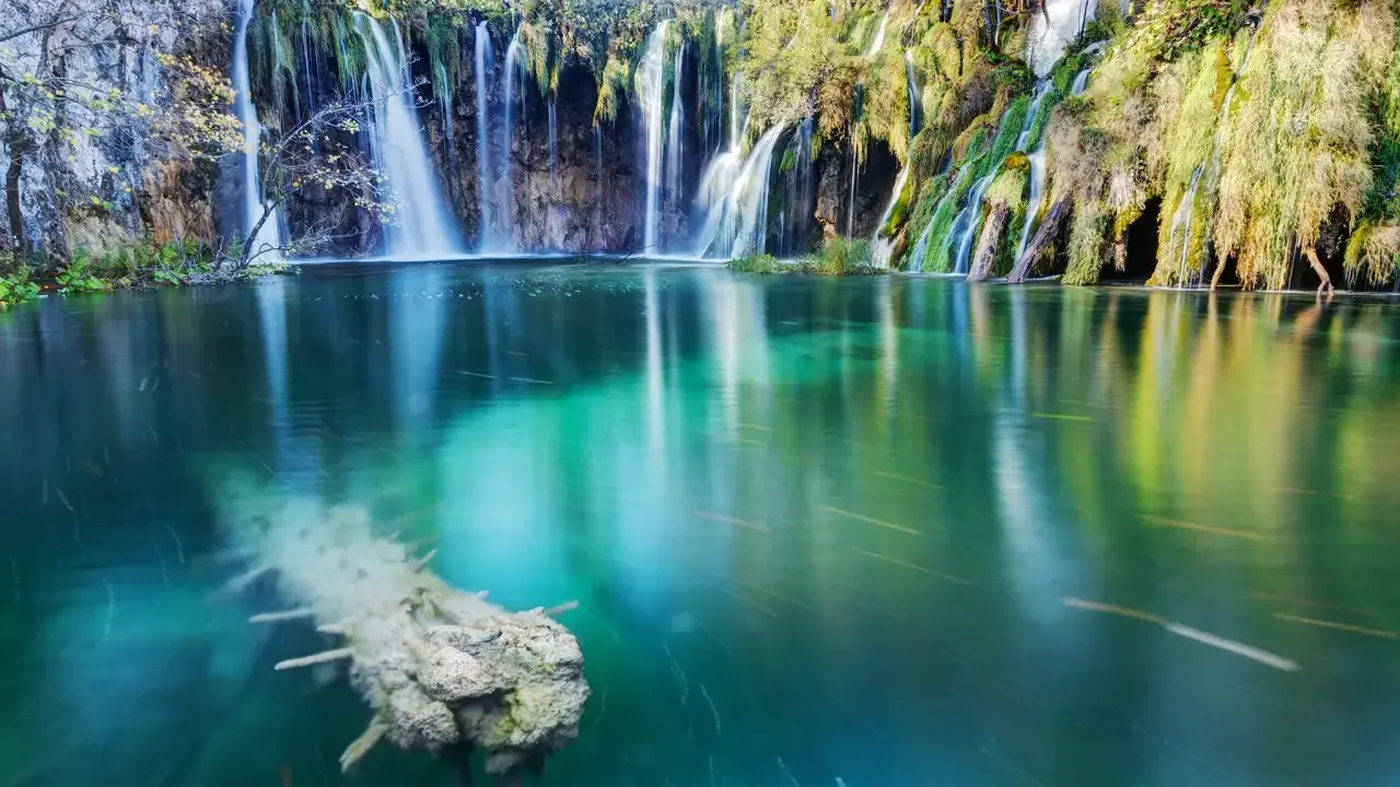 Cascata con effetto grandangolo, acqua turchese e un tronco sommerso.