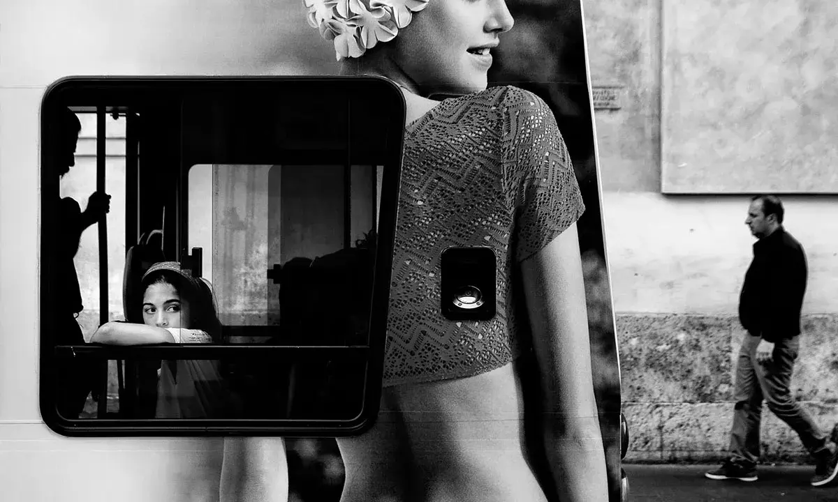 Un uomo cammina mentre una giovane donna guarda fuori da un finestrino. Un'immagine che evoca i grandi fotografi contemporanei.