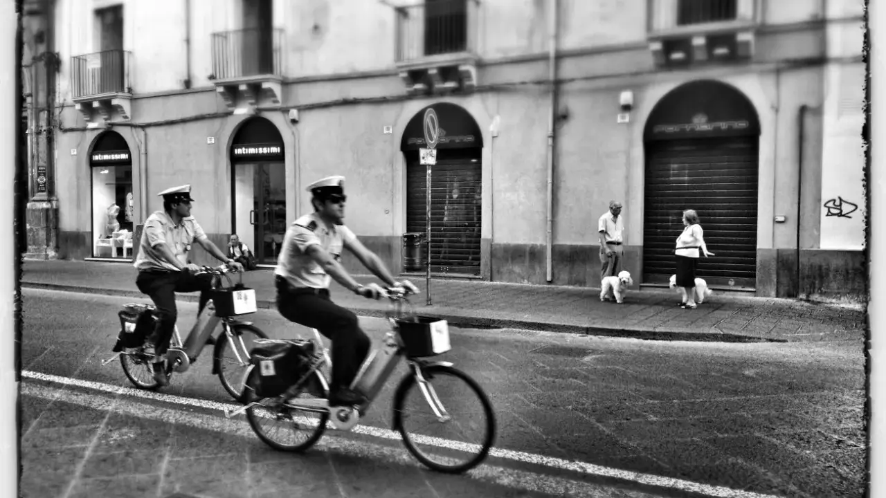 Due agenti in bicicletta pattugliano una strada italiana, mentre una coppia passeggia con i cani sul marciapiede. Italia street photography.