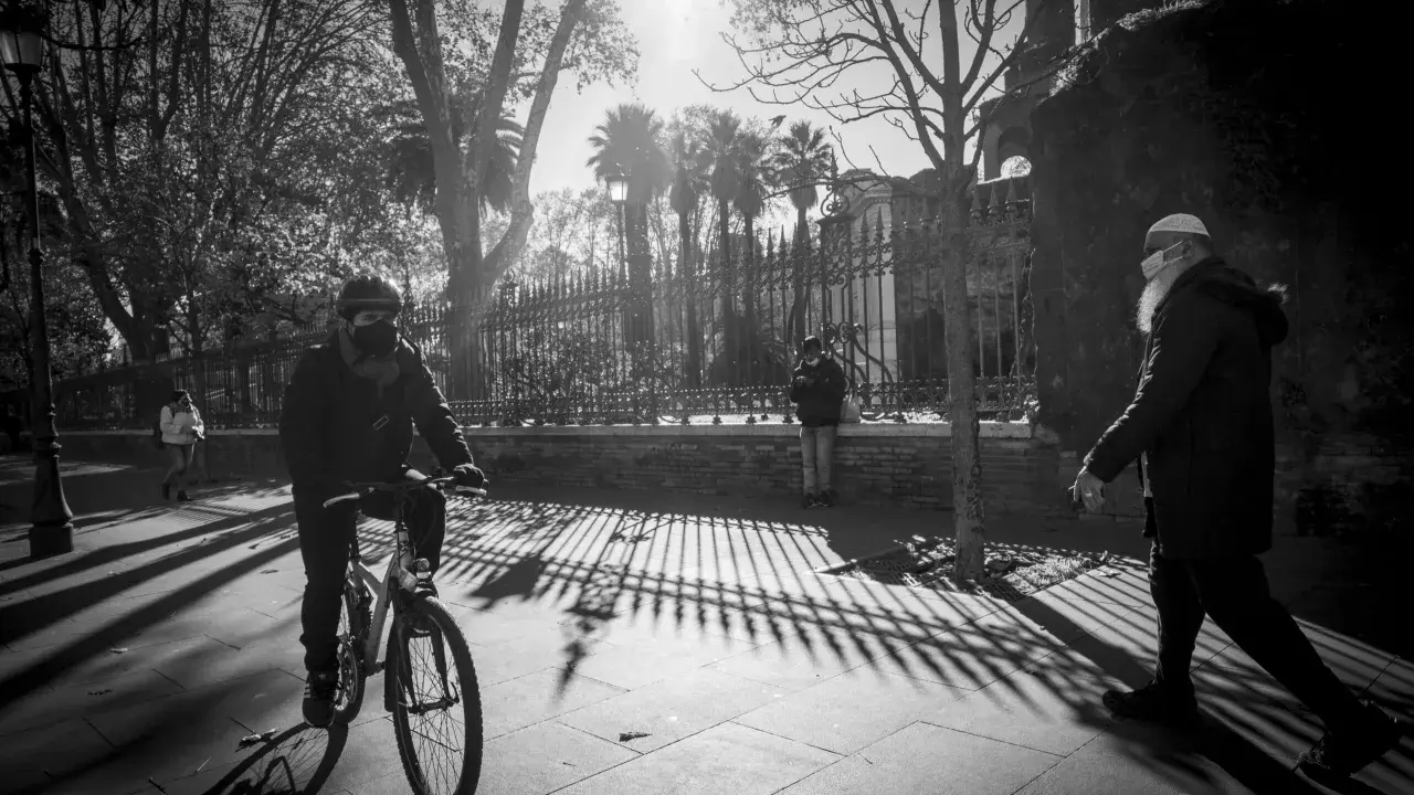 Ombre lunghe e un ciclista mascherato, in uno stile che ricorda robert doisneau, passeggiano sotto il sole.