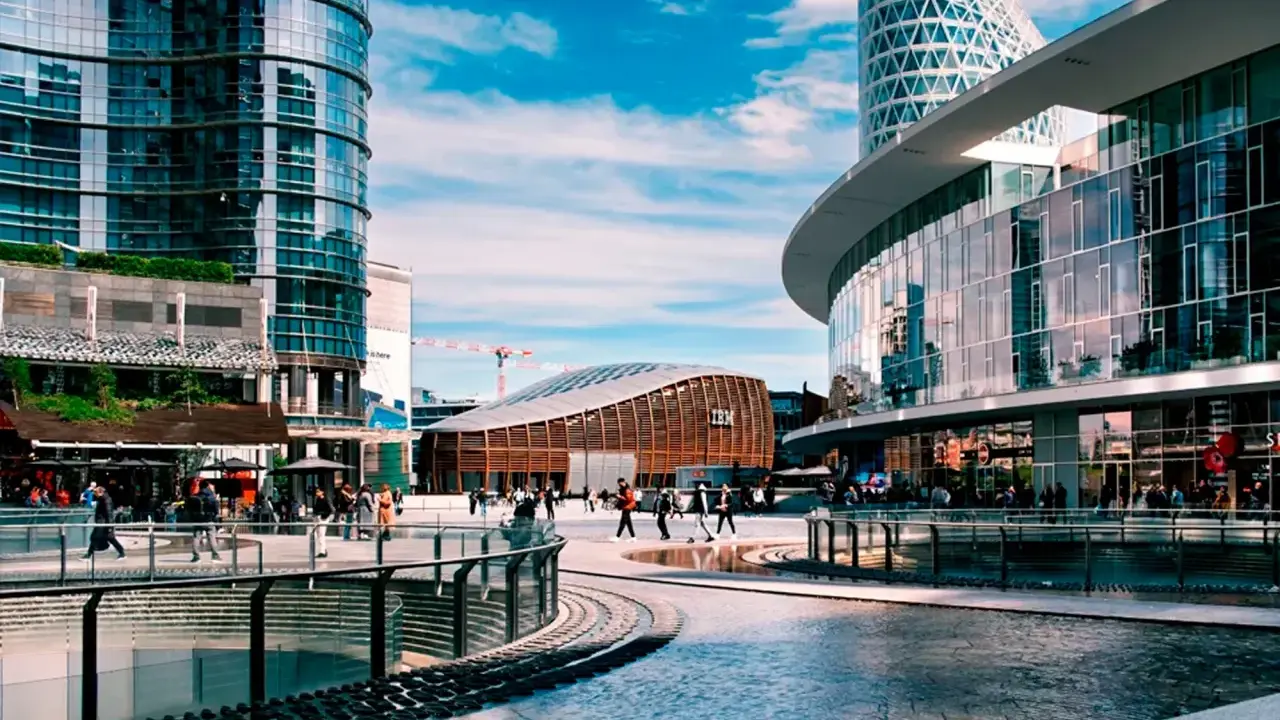 Un'area urbana moderna con grattacieli di vetro, un edificio curvo con il logo IBM e persone che passeggiano. Street photography esempi di vita cittadina.