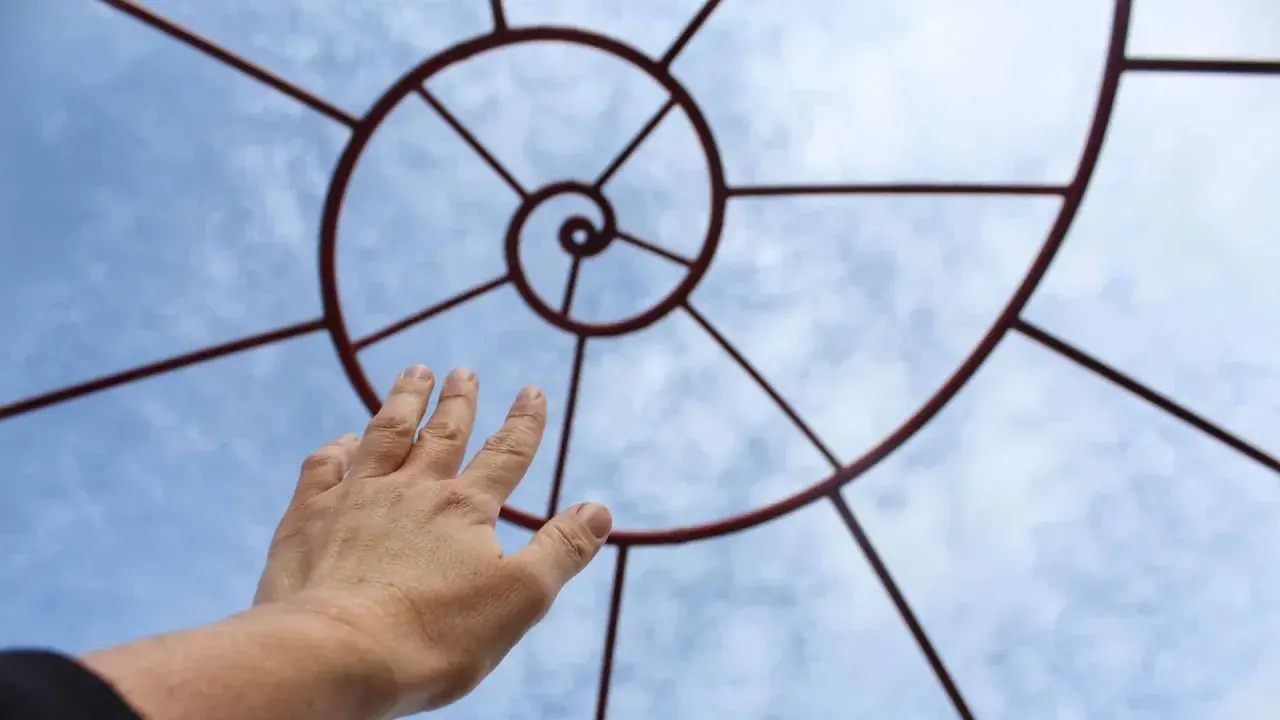Mano tesa verso una spirale metallica che ricorda la composizione aurea, contro un cielo azzurro con nuvole.