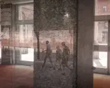 Un riflesso su vetro mostra tre figure che camminano, con un muro di mattoni e un edificio sullo sfondo.