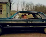 William Eggleston foto: due ragazzi guardano fuori da un'auto nera lucida, con una casa verde sullo sfondo.