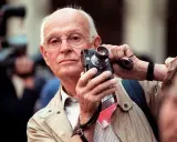 Un anziano fotografo francese famoso con occhiali tiene una vecchia macchina fotografica, pronto a catturare un momento.