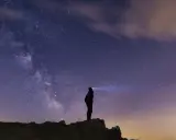 Silhouette di una persona su una collina, che guarda la Via Lattea. Un faro illumina il cielo stellato, un invito a scoprire la Via Lattea.