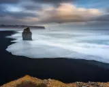 Scogliera solitaria su spiaggia nera, onde bianche in movimento con messa a fuoco infinito. Cielo drammatico.