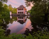 Castello bianco e arancione con torrette, riflessi nell'acqua calma al tramonto, circondato da alberi verdi.