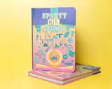 Come fotografare un libro: due quaderni "Sporty Girl Journals" con copertine colorate e illustrazioni sportive su sfondo giallo.