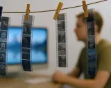 Strisce di pellicola con foto digitali come si fa, appese con mollette. Un ragazzo sfocato osserva.