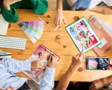 Un team creativo discute idee su un tavolo di legno, con campioni di colore, tastiera, quaderno e foto. Questo è un esempio di come una foto formato JPG può catturare un momento di collaborazione.