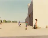 Ghirri fotografo cattura un momento estivo: due bambine e una donna sulla spiaggia, con capanne verdi sullo sfondo e un muro bianco imponente.