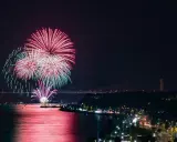 Spettacolare foto fuochi d'artificio rossi e verdi illuminano il cielo notturno sopra un fiume e una città con ponti illuminati.