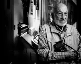 Ritratto di Gianni Berengo Gardin, con la sua inseparabile macchina fotografica al collo, sguardo intenso.