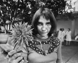 Letizia Battaglia giovane, con uno sguardo intenso, tiene un fiore. Sullo sfondo, figure sfocate in un cortile.
