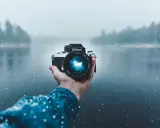 Mano che tiene una fotocamera Nikon in una giornata nevosa, con un lago e alberi sfocati sullo sfondo. Una foto raw per catturare l'essenza dell'inverno.