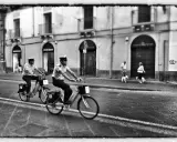 Due agenti in bicicletta pattugliano una strada italiana, mentre una coppia passeggia con i cani sul marciapiede. Italia street photography.