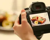 Mano che tiene una fotocamera per fare food photography di un piatto invitante con limone e erbe aromatiche.