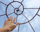 Mano tesa verso una spirale metallica che ricorda la composizione aurea, contro un cielo azzurro con nuvole.