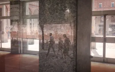 Un riflesso su vetro mostra tre figure che camminano, con un muro di mattoni e un edificio sullo sfondo.