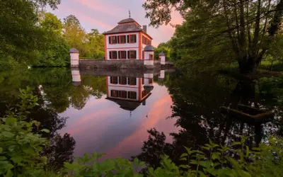 Castello bianco e arancione con torrette, riflessi nell'acqua calma al tramonto, circondato da alberi verdi.