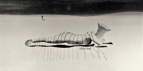 Un uomo solitario osserva una figura distesa, un'opera di un **fotografo surrealista**, con un vaso decorato al posto della testa.