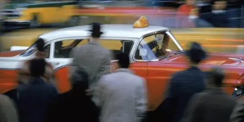 Un taxi rosso e bianco sfreccia in una strada trafficata, un'istantanea che cattura l'essenza di quando nasce la fotografia a colori.