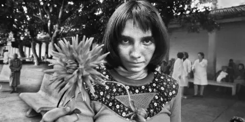 Letizia Battaglia giovane, con uno sguardo intenso, tiene un fiore. Sullo sfondo, figure sfocate in un cortile.