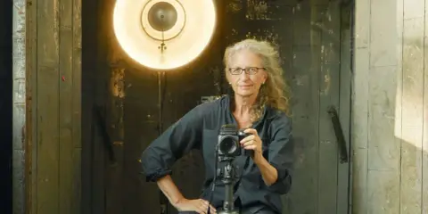 Annie Leibovitz foto: la celebre fotografa con la sua macchina fotografica, illuminata da un grande riflettore.