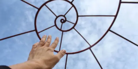 Mano tesa verso una spirale metallica che ricorda la composizione aurea, contro un cielo azzurro con nuvole.
