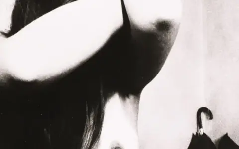 Un corpo femminile in primo piano, con le braccia sollevate, in uno stile che ricorda Bill Brandt. Un ombrello nero è appeso sullo sfondo.