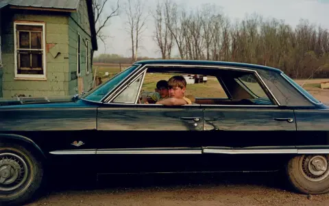 William Eggleston foto: due ragazzi guardano fuori da un'auto nera lucida, con una casa verde sullo sfondo.