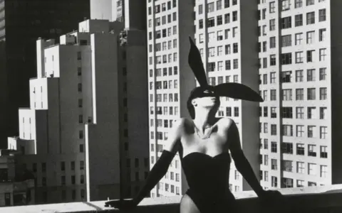 Iconica immagine di Helmut Newton: una donna con orecchie da coniglio e un corsetto nero posa su un tetto, con grattacieli sullo sfondo.